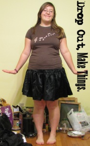 A brown haired girl is wearing a brown shirt with pink music notes and a shiny black knee length skirt